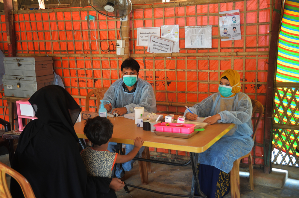 Le personnel d'une organisation humanitaire médicale porte des équipements de protection et des masques faciaux pendant qu'il consulte une mère qui tient son enfant sur ses genoux. Savoir négocier est crucial pour les professionnels de l'humanitaire afin de fournir une attention médicale de qualité.