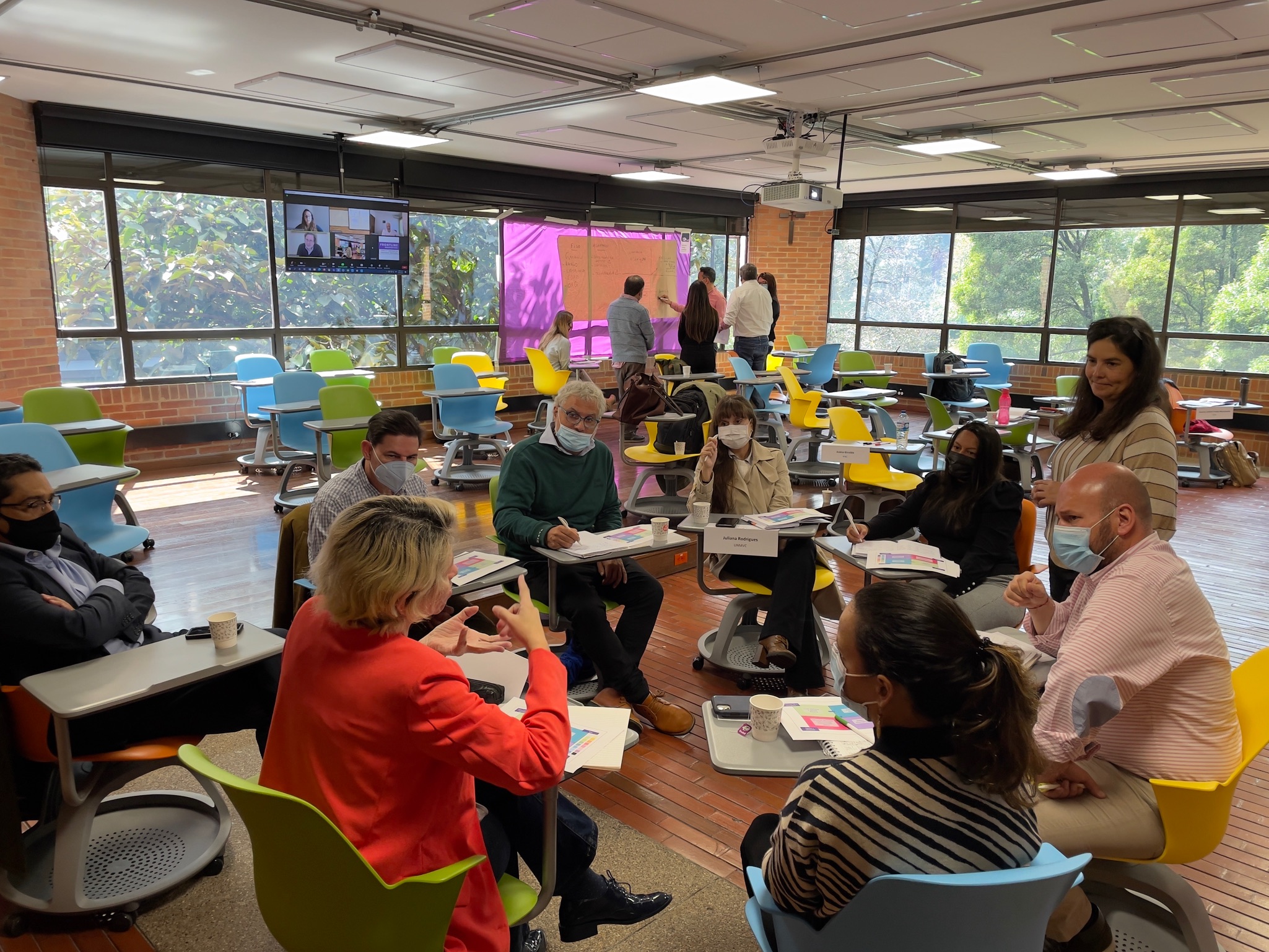 Un grupo de personas están sentadas en círculo hablando sobre cómo ser mejores negociadores humanitarios mientras una facilitadora escucha su conversación. Al fondo, otro grupo de personas debate sobre cómo mejorar su competencias de negociación.